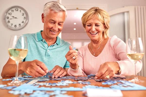 Senior Retired Couple Sitting At Table At Home With Glass Of Wine Doing Jigsaw Puzzle