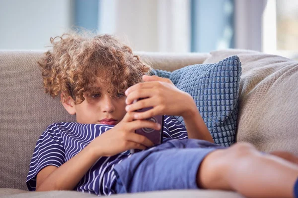 男の子は自宅でソファに横たわっ携帯電話でオンラインゲームをプレイ — ストック写真