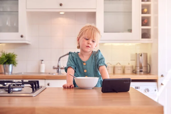 小女孩在家厨房柜台边看手机边吃碗里的早餐谷类食品 — 图库照片
