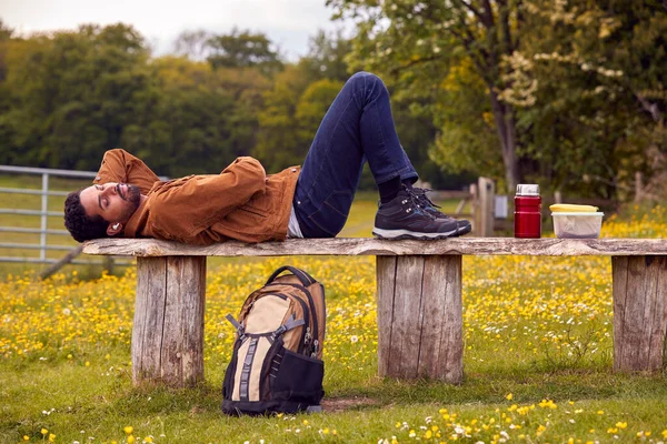 田舎のベンチに寝そべって音楽を聴いたり ワイヤレスイヤホンでポッドキャストを聴いたりする男 — ストック写真