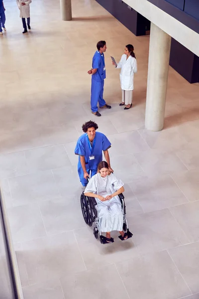 护士头戴刷子在轮椅上推送女病人通过医院大楼的头顶镜头 — 图库照片