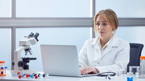 女性实验室工作者穿着实验室工作服用笔记本电脑和显微镜分析实验室血样 — 图库照片