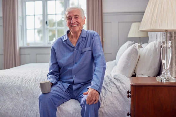自宅でシニア男はお茶の朝のカップを持ってベッドの上に座ってパジャマを着て — ストック写真