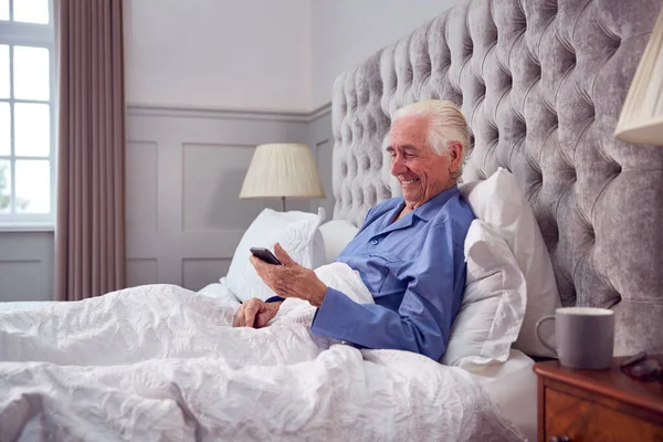 Smiling Senior Man Home Wearing Pyjamas Bed Using Mobile Phone — Foto Stock