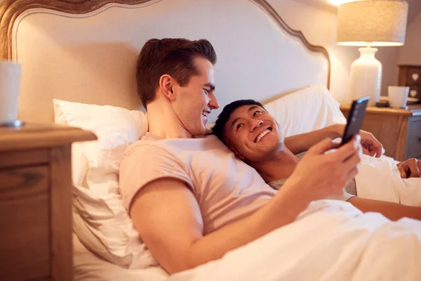 自宅でベッドに横たわっ同じセックス男性カップルを愛します携帯電話でソーシャルメディアを見て — ストック写真