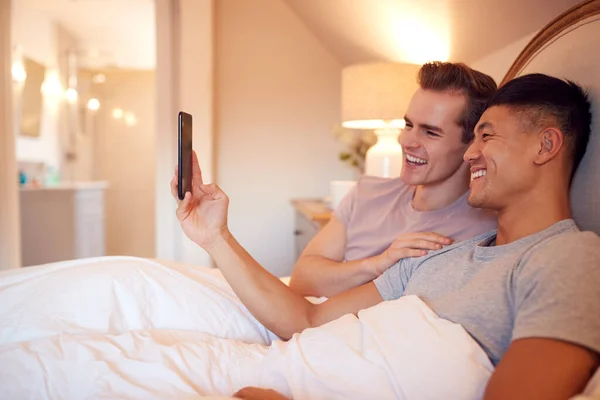 自宅でベッドに横たわっ同じセックス男性カップルを愛します携帯電話でソーシャルメディアを見て — ストック写真