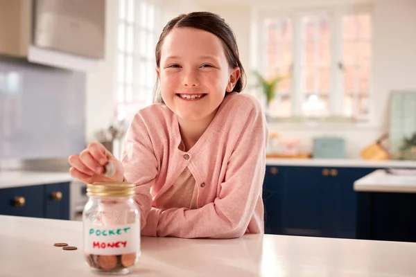 Portrait Of Girl Saving Money Into Jar Labelled Pocket Money On Kitchen Counter At Home