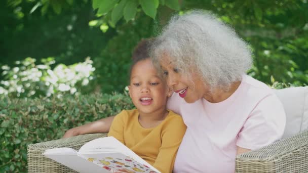 Granddaughter Reading Book Grandmother Sitting Outdoor Seating Garden Home Shot — Vídeo de stock