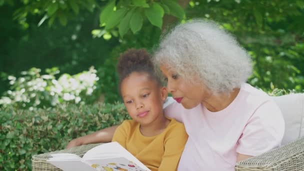 Granddaughter Reading Book Grandmother Sitting Outdoor Seating Garden Home Shot — Stockvideo