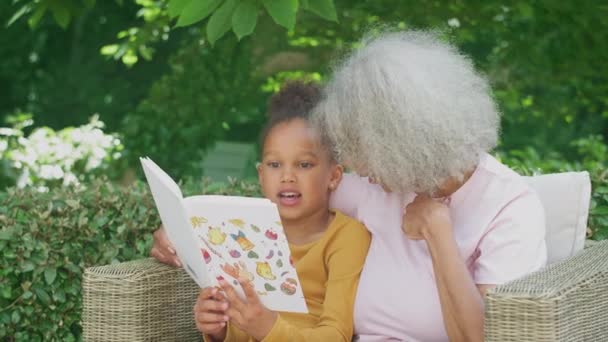 Granddaughter Reading Book Grandmother Sitting Outdoor Seating Garden Home Shot — Stockvideo