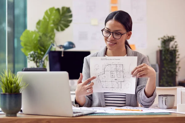 Female Architect In Office Sitting At Desk Showing Plans For New Building On Video Call