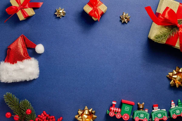 Composizione natalizia. Un trenino di legno, un albero di Natale con bacche rosse, fiocchi d'oro, regali, un cappello da Babbo Natale su sfondo blu scuro. Capodanno. Layout piatto, vista dall'alto, spazio di copia — Foto Stock