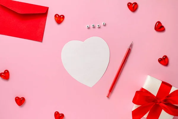 The concept of Valentines Day. A red envelope, hearts, a white sheet for a note, a pen, the word love, a gift with a satin ribbon on a pink background. Flat lay, copy space — Fotografia de Stock