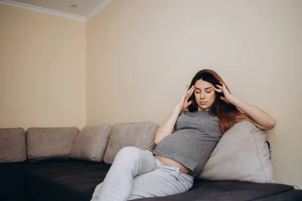 Pregnant woman with hand on forehead suffering headache sitting on a couch in the living room at home