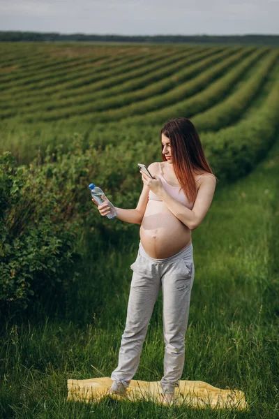 美丽的年轻孕妇拿着一瓶水在大自然的电话里交谈 在绿色的夏季田野的背景下 — 图库照片