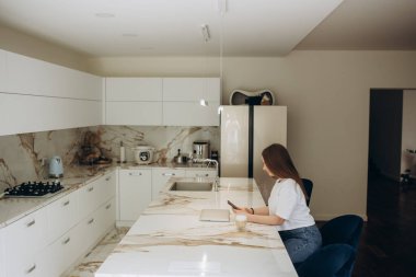 Genç ve güzel bir kadın cep telefonu kullanıyor ve mutfakta kahve içiyor.