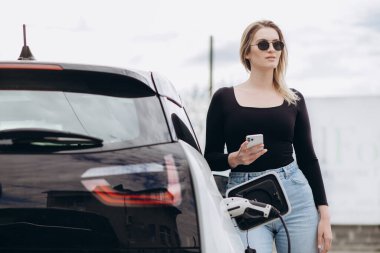 Telefon kullanarak sohbet ediyorlar. Elektrikli arabalardaki kadın gündüz şarj istasyonunda. Yepyeni bir araç..