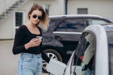 Telefon kullanarak sohbet ediyorlar. Elektrikli arabalardaki kadın gündüz şarj istasyonunda. Yepyeni bir araç..