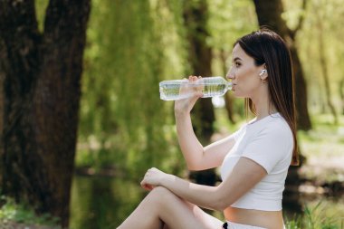 Spor elbiseli, spor elbiseli, spor yaparken su içen, sonbahar güneşli bir günde parkta yoga yapan bir kadın..