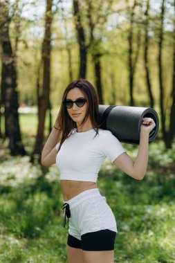 Spor kıyafetli sıska bir kadın yoga minderini tutuyor ve dışarıda dikilip başka tarafa bakıyor. Parkta güneşli bir günde çekici beyaz kadın sporcu. Spor, spor, aktif yaşam tarzı konsepti.