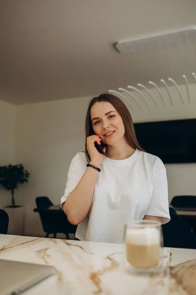 Talks by the phone. Young european woman is indoors at kitchen indoors with healthy food