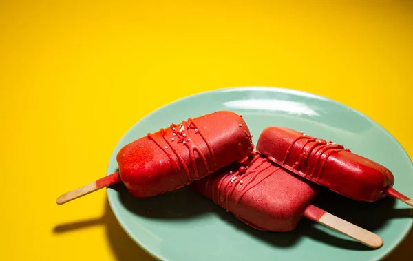 Cake in the form of ice cream on a stick among flowers, close-up. Cheesecake on a stick. Ice cream in glaze. Yellow background, top view