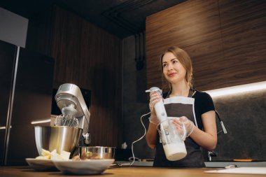 Önlük giymiş güzel bir kadın pastacı modern mutfakta kremayla karıştırıcıyı yener.