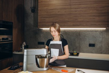 Genç bir kadın mutfakta elmalı turta pişiriyor önlüğüyle tezgahın başında duruyor bardaktaki taze malzemeleri çırpmak için bir karıştırıcı kullanıyor. Önünde elmalar, yumurtalar ve pişirme tenekesi var.