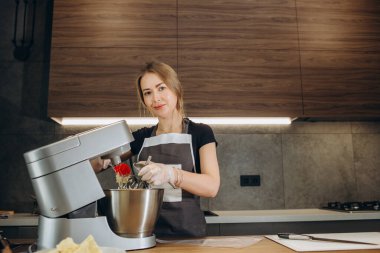 Genç bir kadın mutfakta elmalı turta pişiriyor önlüğüyle tezgahın başında duruyor bardaktaki taze malzemeleri çırpmak için bir karıştırıcı kullanıyor. Önünde elmalar, yumurtalar ve pişirme tenekesi var.