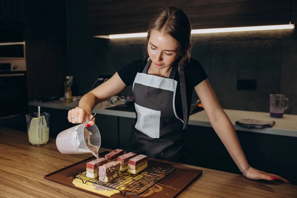 Smiling female confectioner pouring liquid chocolate gommage on biscuit cake in metal form working at catering service restaurant. Woman chef in uniform at assembling mousse dessert process