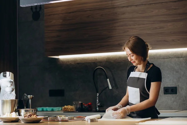 Beautiful female confectioner prepares sweet in modern kitchen looking at dough