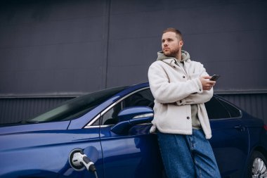 Adam arabanın kaputuna oturmuştu. Arabası şarj istasyonunda şarj oluyor. Bir adam akıllı telefon ekranına bakar ve gülümser..