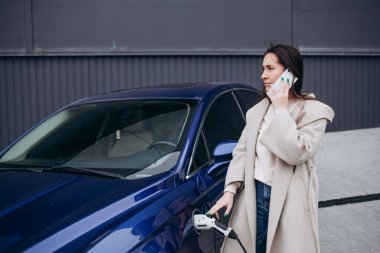 Modern kablosuz teknolojiler. Telefonla. Elektrikli arabalardaki kadın gündüz şarj istasyonunda. Yepyeni bir araç..