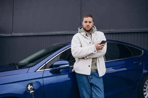 The guy sat down on the hood of the car. His car is charging at the charging station. A man looks at the smartphone screen and smiles.