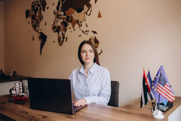 Concept for travel agent. Young blonde woman with glasses smiling, looking at camera and using laptop. Travel agency office interior with big world map and window