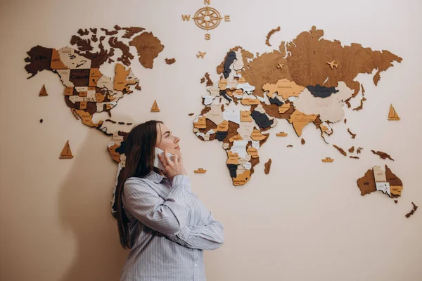 Photo of female travel agent. Young woman talking on phone, smiling and looking at camera. Travel agency office interior with big world map
