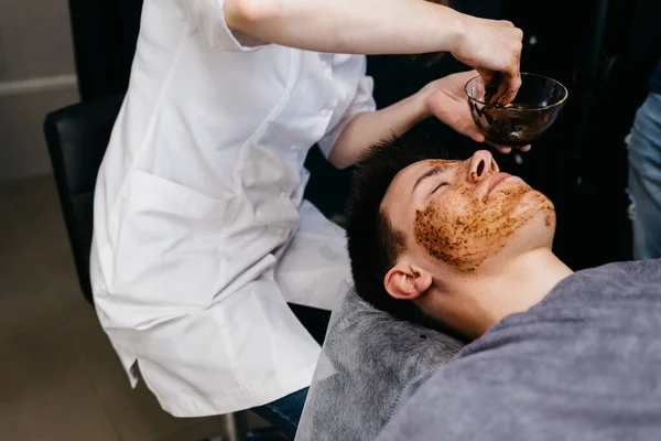 Photo of cheerful Caucasian man with coffee scrub mask on face, touches forehead and chin, smiles broadly, shows white perfect teeth, has muscular body, models in studio agaisnt blue background