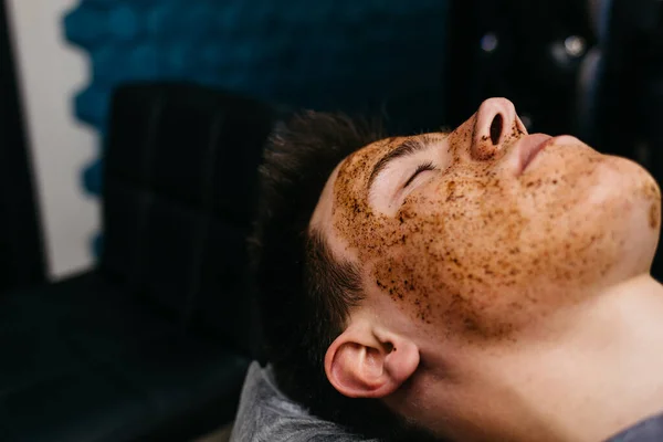 Photo of cheerful Caucasian man with coffee scrub mask on face, touches forehead and chin, smiles broadly, shows white perfect teeth, has muscular body, models in studio agaisnt blue background