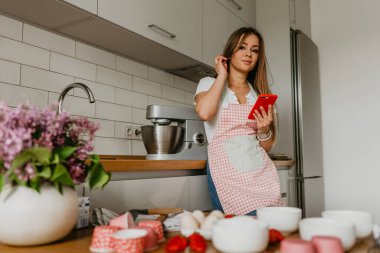 Sebzeli mutfakta cep telefonu kullanırken gülümseyen genç kadın