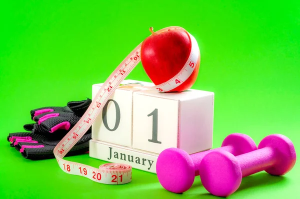 New year resolution, start a diet and stop procrastinating concept with an apple, pink dumbbells and gym gloves next to a cube calendar showing the date of january 1st on bright vivid green background