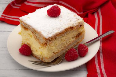 Bir parça vanilyalı ve kremalı pasta (Almanca: Cremeschnitte, Lehçe: Napoleonka veya Kremowka) ve ahududu beyaz ahşap arka planda ve kırmızı çay bezinde.