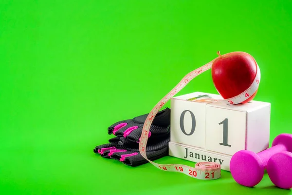 New year resolution and start a diet concept with an apple, pink dumbbells and gym gloves next to a cube calendar showing the date of january 1st on bright vivid green background with copy space