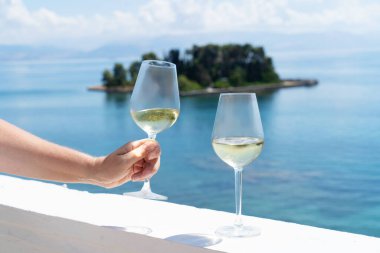 Arka planda Corfu deniz manzaralı beyaz şarap kadehlerinden birini tutarken.