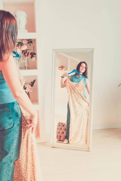 Joven morena de pie frente al espejo en casa sosteniendo vestido de noche en lentejuelas de oro —  Fotos de Stock