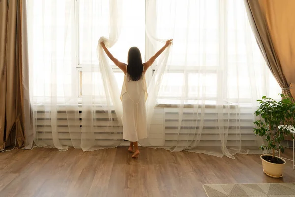 Mujer joven se para junto a una gran ventana en vestido blanco de seda después de despertar en casa —  Fotos de Stock