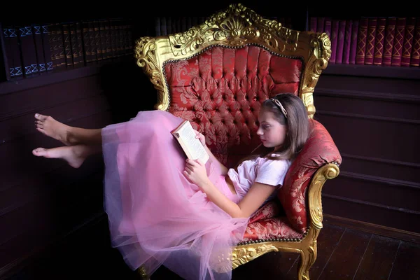 Прекрасна дівчина-підліток в рожевій спідниці читає книгу, сидячи збоку на шикарному золотому кріслі — стокове фото