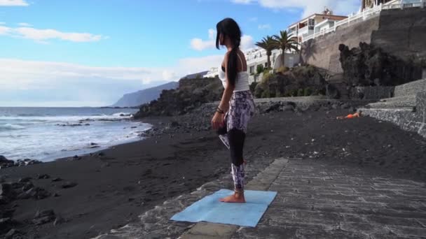 Жінка в спортивному одязі практикує йогу і медитує на пляжі вранці — стокове відео