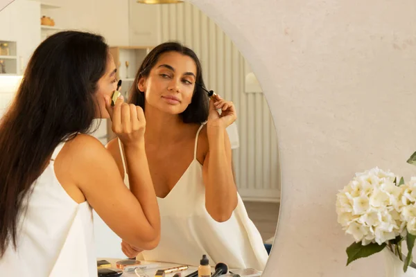 Young beautiful woman with long straight dark hair and fresh clean skin doing morning makeup routine, using eye mascara in front of mirror table at home. Beauty and facial cosmetics concept