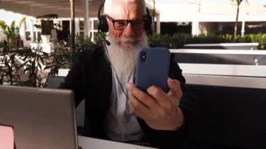 Senior smiling man working with laptop and talking via mobile phone on cafe or home terrace