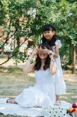 Küçük kız annesiyle eğleniyor, avuçlarıyla gözlerini kapatıyor. İkisi de beyaz elbiseler giyip parkta piknik yapıyorlar. Ağaç gölgesinde düşüşün son sıcak günlerinin tadını çıkarıyorlar..